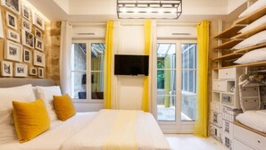 A bedroom with a bed, a flat-screen TV, a window with yellow curtains, and a bedside table with a lamp.
