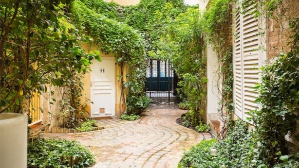 A narrow alleyway with a cobblestone path, flanked by lush greenery and buildings with shutters.