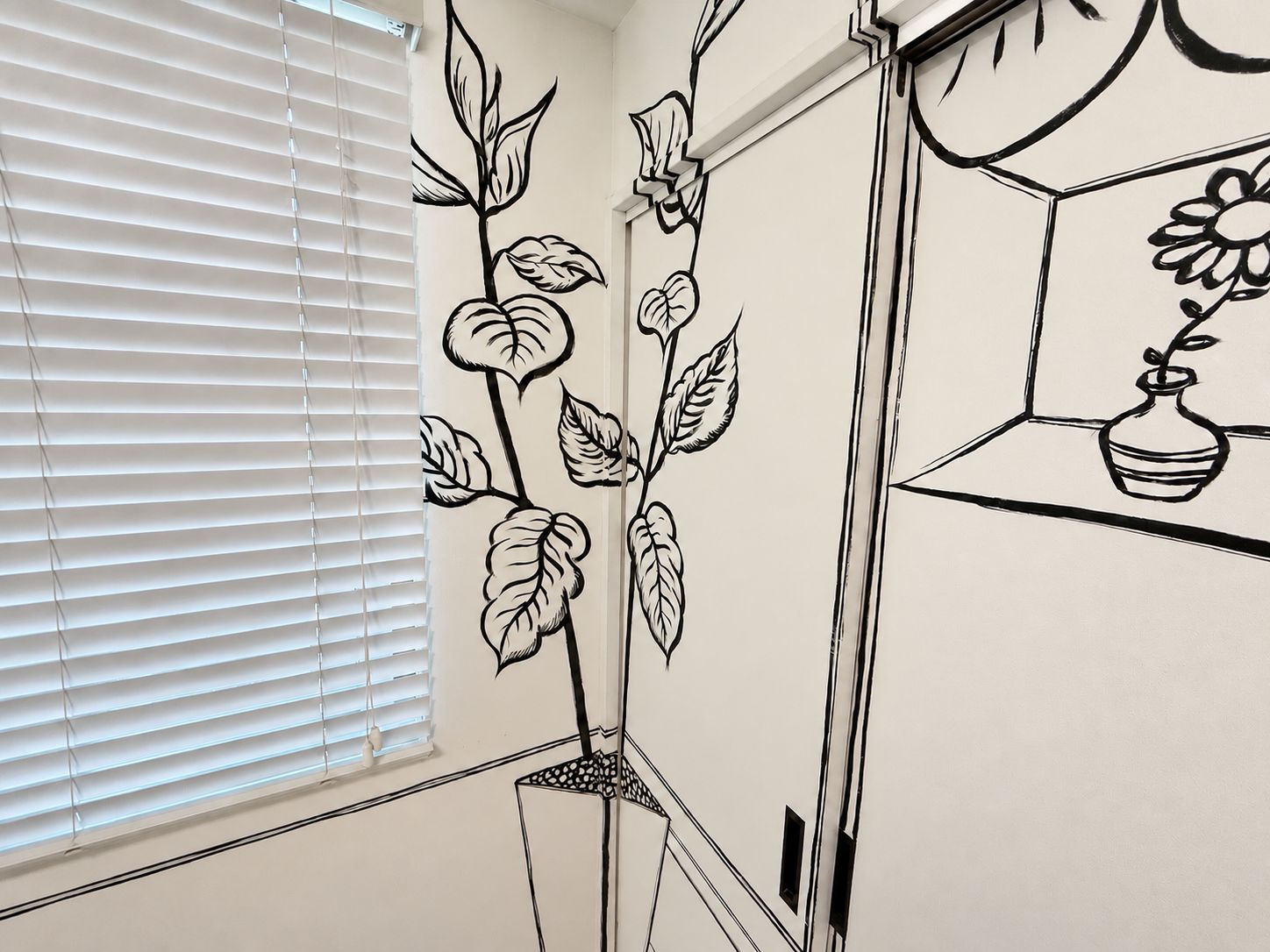 Una habitación con ventana y persianas, una pared con un mural floral y un estante pequeño con un jarrón.