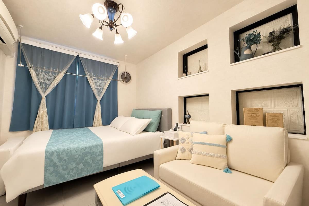 A hotel room with a bed, a sofa, a desk with a lamp, a ceiling fan, and framed pictures on the wall.