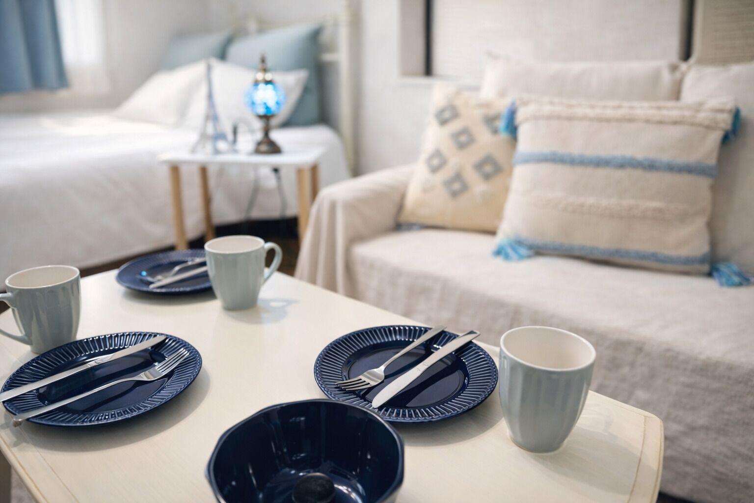 A neatly arranged breakfast table with plates, cutlery, and cups, set against a backdrop of a bed and a nightstand with a lamp.