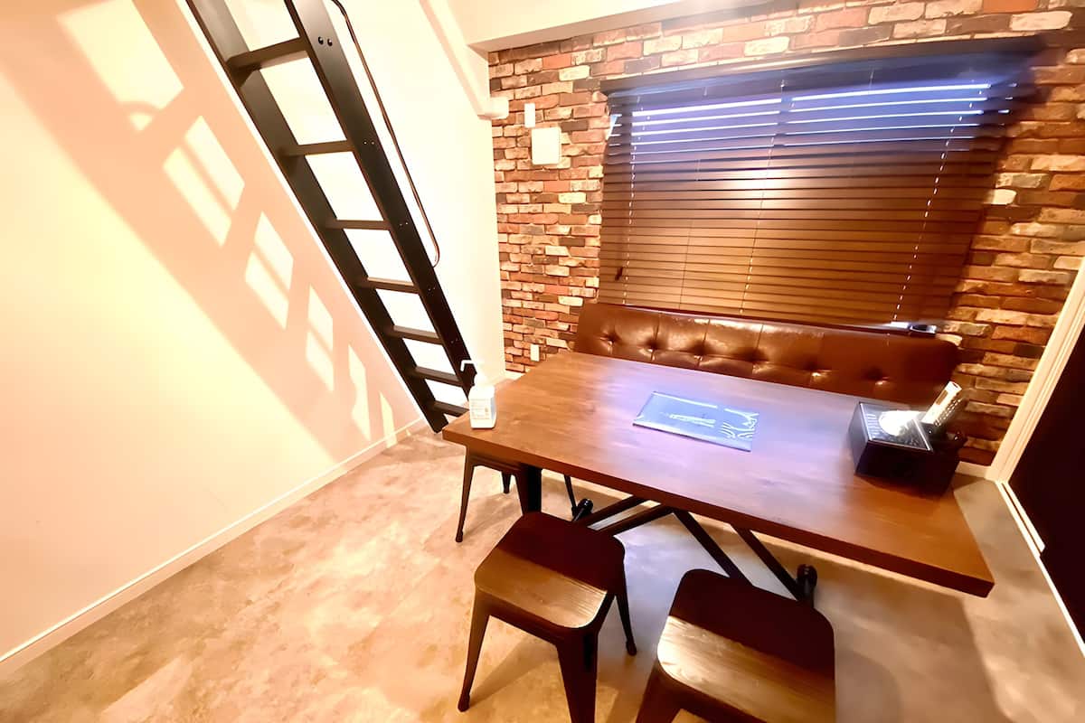 A dining area with a wooden table, two chairs, a bench, and a brick wall with a window.