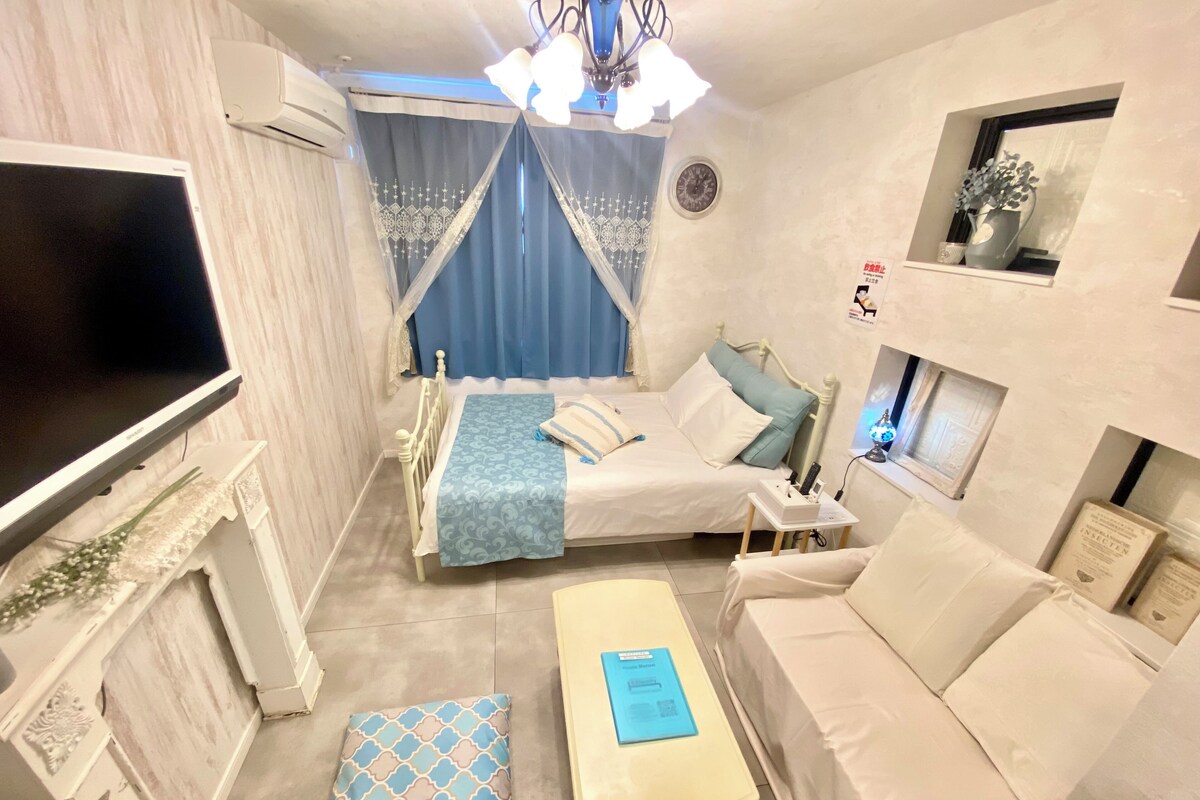 A compact hotel room with a bed, a sofa, a small table, a TV, and a window with a clock.