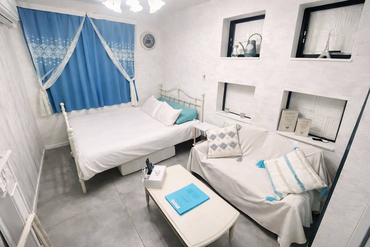 A bedroom with a bed, a smaller bed, a bedside table, a window with curtains, a wall clock, and a shelf with decorative items.