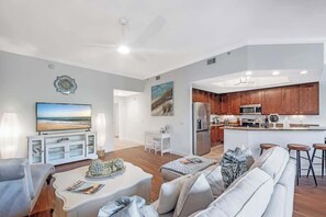 A modern living room with a TV, white furniture, and a kitchen with wooden cabinets.