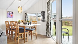 Un espace repas avec une table blanche et des chaises en bois, un luminaire suspendu et une vue sur l’extérieur grâce à de grandes fenêtres.