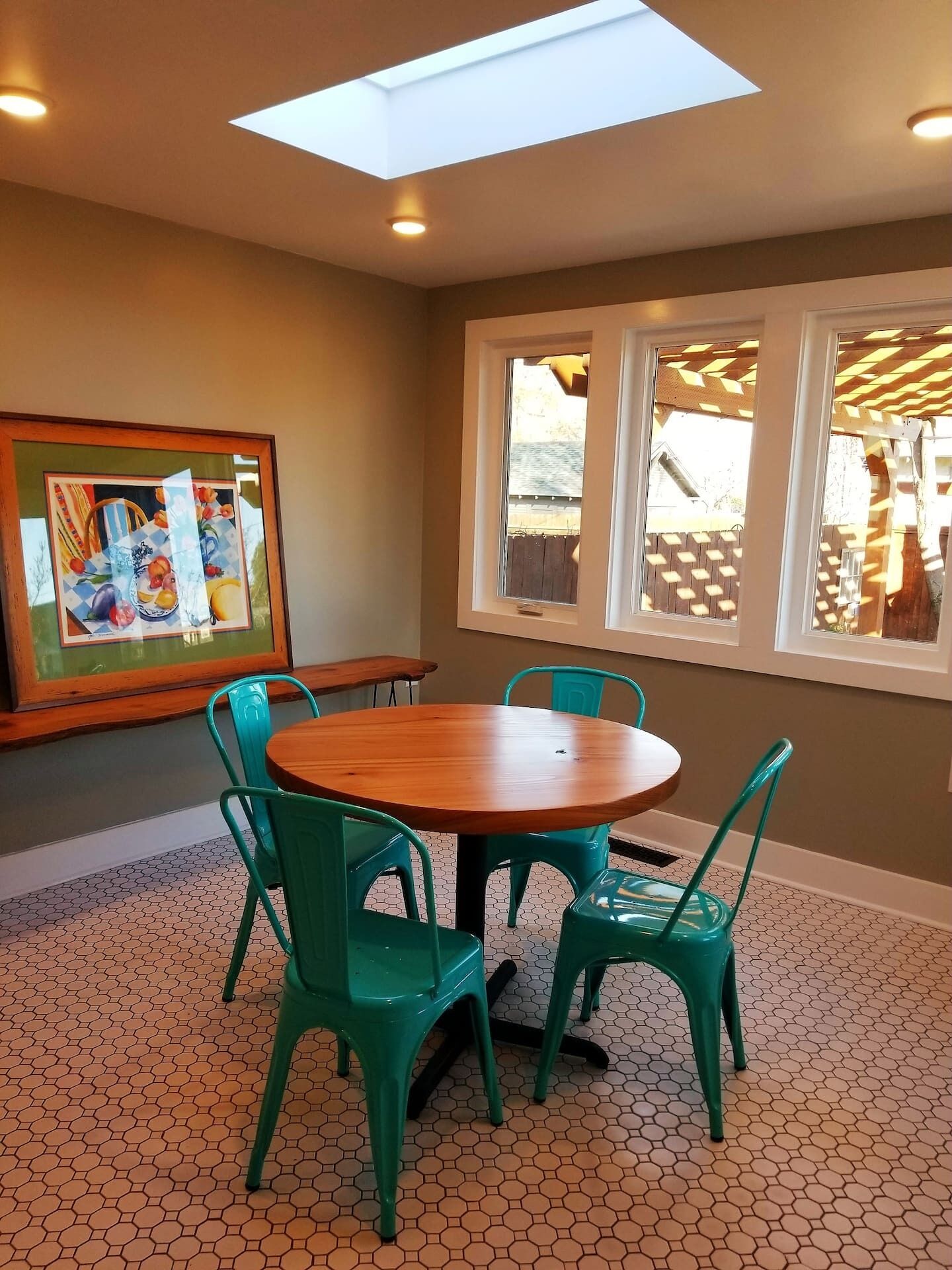 A dining area with a round wooden table, four green chairs, a hexagonal patterned floor, a painting on the wall, and a window with a view of a backyard.