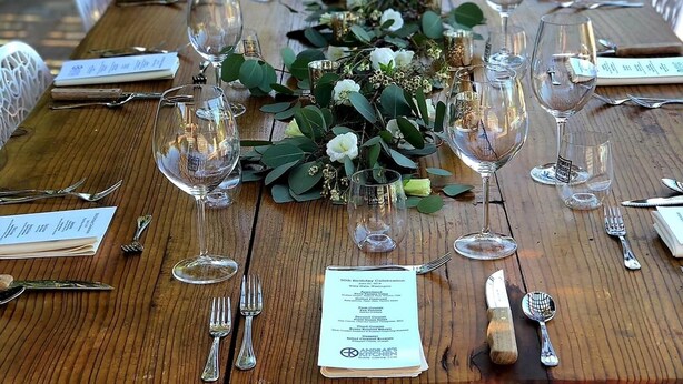 A long wooden table set for a formal outdoor event with name cards, wine glasses, and cutlery.