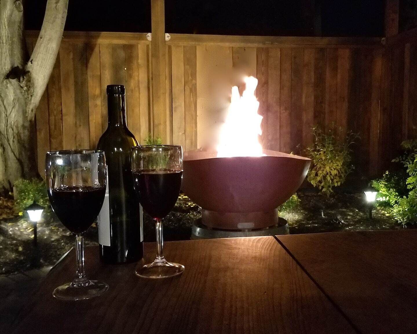 A wooden table with two wine glasses and a bottle, a fire pit with flames, and a wooden fence in the background.