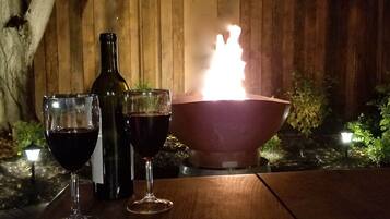 A wooden table with two wine glasses and a bottle, a fire pit with flames, and a wooden fence in the background.
