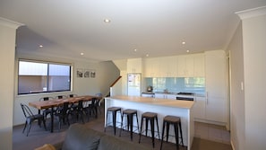 A modern kitchen with white cabinets, a central island with stools, a dining area with a wooden table and chairs, and a large window.