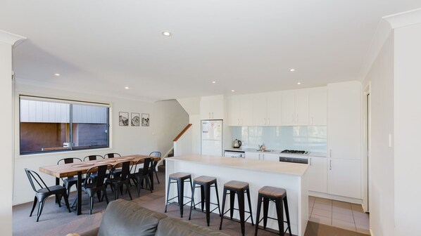 Una cocina moderna con armarios blancos, una isla central con taburetes y una zona de comedor con mesa y sillas de madera.
