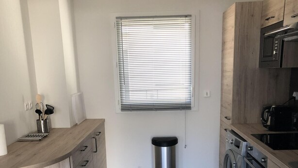 A modern kitchen with wooden cabinets, a stainless steel trash bin, a built-in washing machine, and a window with blinds. - Rental house in Valras plage (34) 6/8 people (Sérignan)
