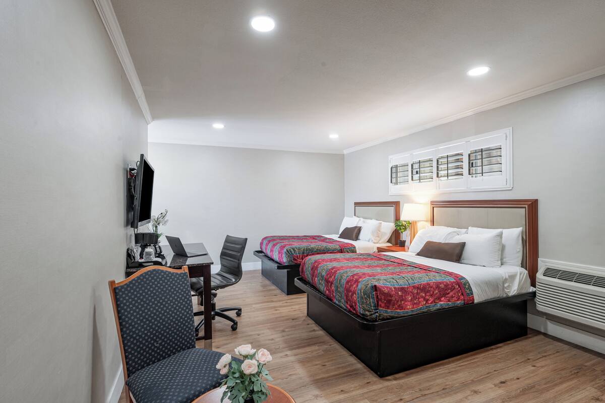 A hotel room with two beds, a desk, a chair, a TV, and a window with blinds.