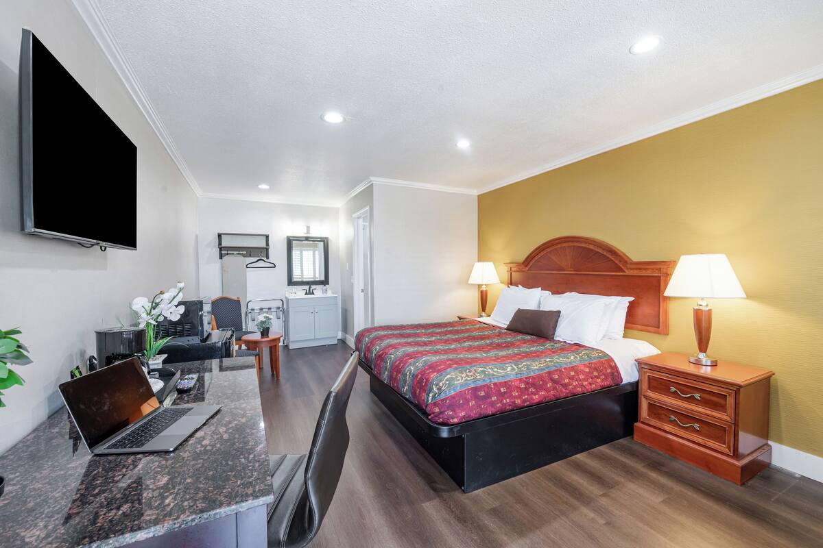 A hotel room with a bed, bedside lamps, a desk with a laptop, a TV, and a small desk area with a chair and a plant.