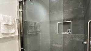 A modern bathroom with a glass-enclosed shower, a white towel rack, and a small shelf. - Ritz Inn San Francisco SFO Airport (San Bruno)