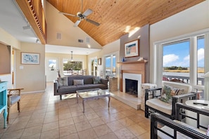 A spacious living room with a fireplace, a ceiling fan, and large windows offering a view of the outdoors.