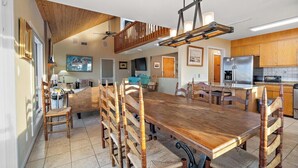 Een ruime eethoek met een lange houten tafel en stoelen, een keuken met houten kasten, en een plafondventilator.