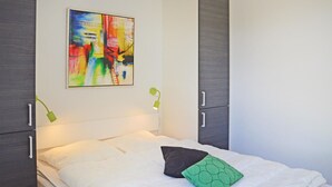 Een net opgemaakt bed met witte lakens en een kussen met groen patroon, een kleurrijk abstract schilderij aan de muur en een groene bureaulamp.