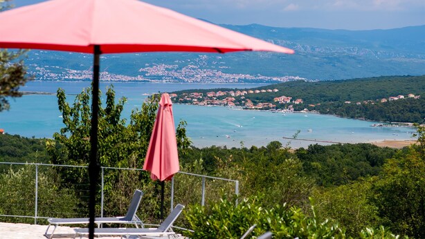 En terrasse med rød parasol, liggestole og et smukt udsigt over en kystby og bjerge.