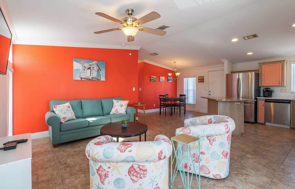 A living room with a teal sofa, a coffee table, and two patterned armchairs. A ceiling fan is visible, and the room has a bright orange accent wall.