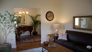 A living room with a wooden coffee table, a black sofa, a lamp, a clock, a potted plant, and a piano in the background. - The Tudor Rose - A classy, comfortable getaway (San Anselmo)