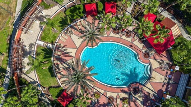 Une vue aérienne d’une piscine circulaire entourée de palmiers et d’ombrelles rouges, avec une allée pavée et une clôture en arrière-plan.