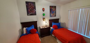 A bedroom with two beds, each with red bedding and blue pillows. There are posters on the wall, a small table with a lamp, and a window with blinds.