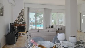 A modern living room with a stone fireplace, a sofa, a dining table with wine glasses, and a view of a pool. - Piccolo Paradiso Villa A (Lachania)