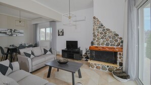 A modern living room with a stone fireplace, a sofa, a coffee table, and a television. - Piccolo Paradiso Villa A (Lachania)