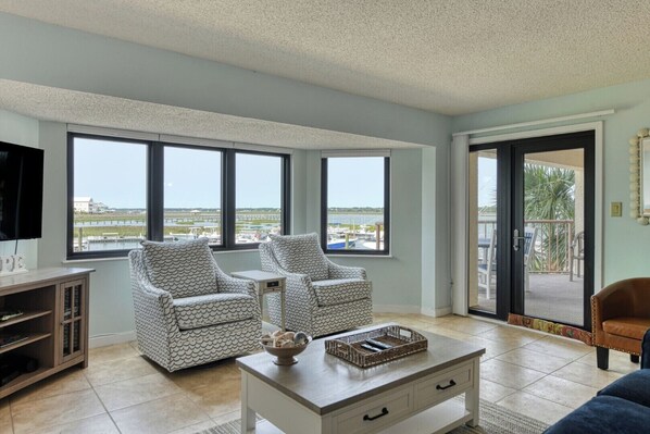 A living room with two patterned armchairs, a coffee table with a basket, a TV on a stand, and large windows with a view of a marina.