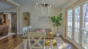 A dining area with a wooden table, white chairs, a chandelier, and a large window with a view of trees. - Alpaca My Bags & Alpaca Experience! (Silt)