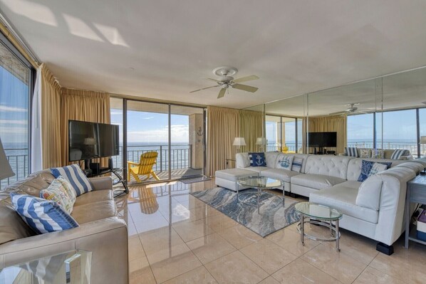 A spacious living room with a large sofa, a coffee table, and a TV. There is a balcony with a chair and curtains, and a ceiling fan.