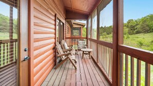 Una terraza de madera con vistas a un paisaje verde, un espacio cubierto con sillas y una mesa, y una puerta con ventanal.