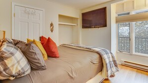 Un dormitorio con cama, almohadas, un televisor montado, una ventana con vistas y una puerta con ganchos.