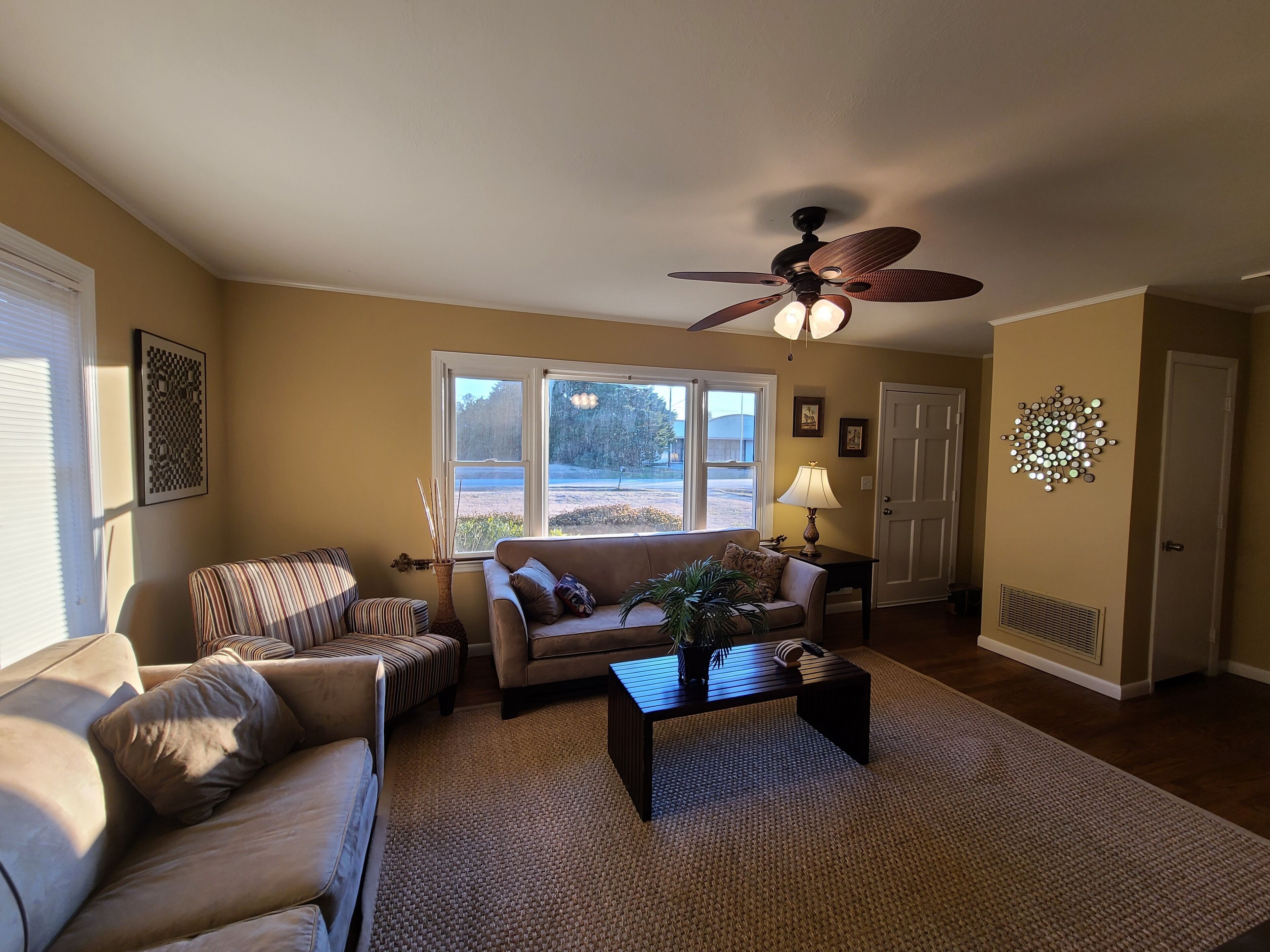 A living room with a sofa, armchair, and coffee table, a ceiling fan, and a large window with a view of the outdoors.