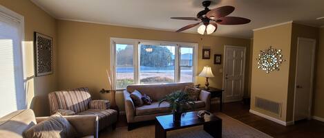 A living room with a sofa, armchair, and coffee table, a ceiling fan, and a large window with a view of the outdoors.