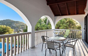 A covered outdoor seating area with a round table and chairs, white railings, and a view of a hillside with trees and buildings. - Beautiful home in Tisno (Tisno)