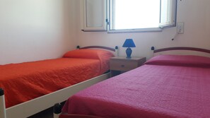 Two single beds with red and pink bedding, a nightstand with a lamp, and a window with a view of the outside.
