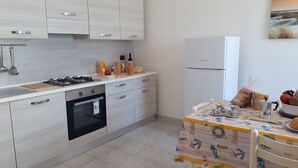 A modern kitchen with a gas stove, oven, and refrigerator, a dining table with a children's table, and a wall-mounted sink. - Central apartment just steps from the beach with balcony (San Foca)