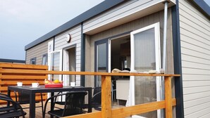 A modern mobile home with a deck area featuring a table and chairs, a basket of fruits, and a wooden railing.