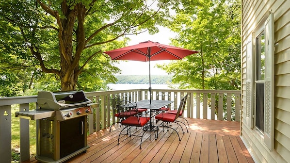A wooden deck with a grill, red chairs, and a table under a red umbrella, overlooking a lake.