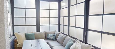 A sunroom with a large window, a wooden table, and a cushioned seating area.
