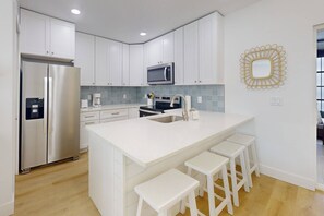 A modern kitchen with white cabinets, a stainless steel refrigerator, a microwave, and a sink. There are three white stools at the island.