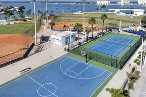 Un court de tennis avec un stand de vente de rafraîchissements, entouré d’une clôture et de palmiers, offrant une vue sur l’océan et les bâtiments en arrière-plan.