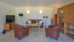 Een woonkamer met een bank, twee fauteuils, een salontafel, een televisie, een vloerlamp en houten planken.