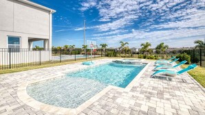 A swimming pool with a basketball hoop, surrounded by a paved patio and a fence.