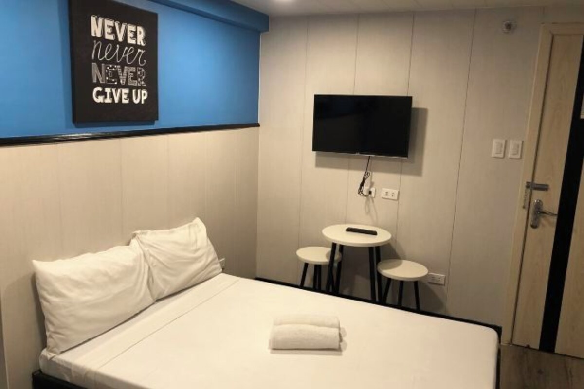 Hotellihuone, jossa on sänky, seinälle asennettu televisio ja pieni pöytä, jonka ympärillä on kaksi tuolia.