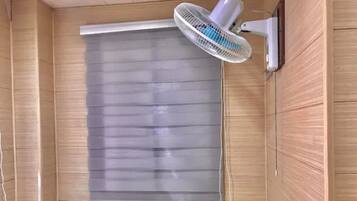 Una habitación con un ventanal grande cubierto por persianas, un ventilador de pared y una pequeña mesa.