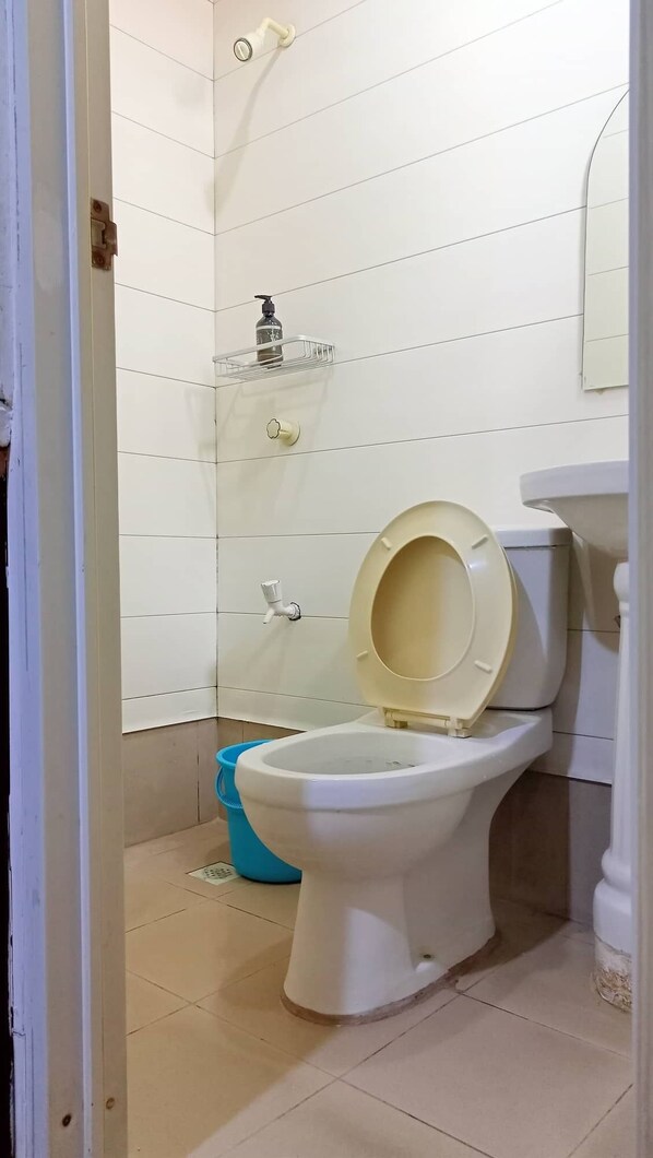 A bathroom with a toilet, a blue bucket, and a soap dispenser on a shelf.